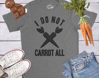 May include: A heather gray short-sleeve t-shirt featuring a black graphic of two crossed carrots and the text I DO NOT CARROT ALL. The shirt is displayed with a brown chocolate rabbit, a bunch of fresh orange carrots, white sneakers, and blue jeans.