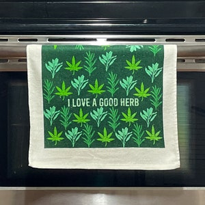 May include: A white kitchen towel with a green background and a pattern of green leaves. The text "I LOVE A GOOD HERB" is printed in white on the towel.