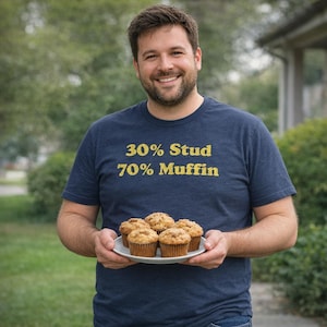 May include: A navy blue short-sleeved t-shirt with the yellow text 30% Stud 70% Muffin printed on the chest. The shirt is worn by a person holding a white plate containing four golden-brown muffins with a textured, crumbly top surface.