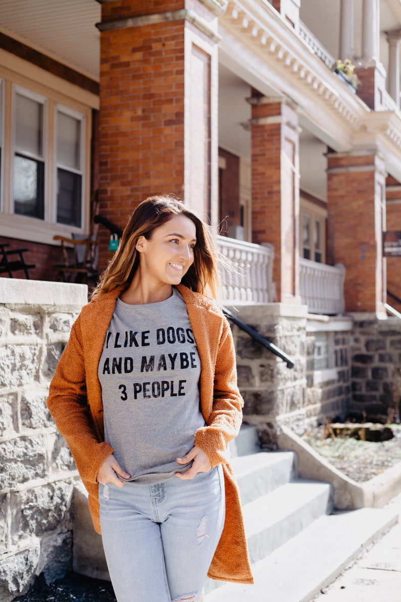 May include: A woman wearing a gray t-shirt with the text "I LIKE DOGS AND MAYBE 3 PEOPLE" and a rust-colored coat. She is standing on a stone staircase in front of a brick building.