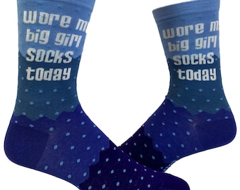 May include: A pair of crew socks featuring a gradient blue color scheme with a polka dot pattern. The text "wore my big girl socks today" is printed in white block letters on the upper portion of each sock against a light blue background.