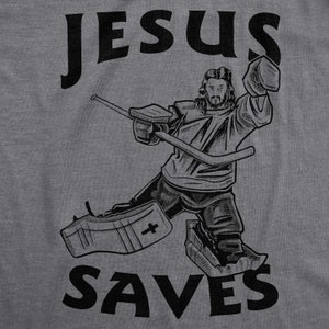 Puede incluir: Una camiseta gris con un gr&aacute;fico negro de Jes&uacute;s jugando al hockey. El gr&aacute;fico muestra a Jes&uacute;s con un uniforme de hockey y sosteniendo un palo de hockey. El texto "JESUS SAVES" est&aacute; impreso en negro debajo del gr&aacute;fico.