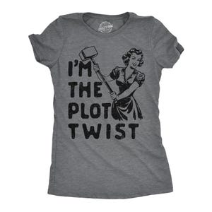 Puede incluir: Camiseta gris con un gráfico negro de una mujer sosteniendo un martillo y el texto "I'm the plot twist".
