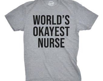 May include: A heather gray short-sleeved t-shirt with a crew neckline. The front of the shirt features the black text "WORLD'S OKAYEST NURSE" printed in a bold, sans-serif font. The garment has a standard fit with a straight hemline.