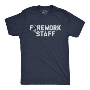 May include: A navy blue short-sleeve t-shirt featuring the white distressed text "FIREWORK STAFF" on the chest, with the letter "I" replaced by a graphic of a firework rocket. The shirt has a crew neckline and a standard fit design.