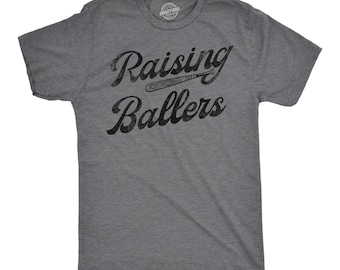 May include: A charcoal grey heather short-sleeve t-shirt with a crew neckline. The front features black distressed text that reads Raising Ballers, with a small graphic of a baseball bat positioned between the two words of the text.