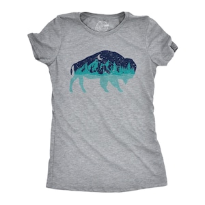 May include: A heather gray short-sleeve t-shirt featuring a graphic of a bison silhouette. The interior of the bison shape displays a night landscape with dark blue mountains, a crescent moon, starry sky, and a light teal forest and water reflection.