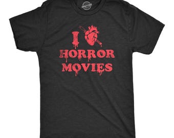 May include: A charcoal grey short-sleeve t-shirt featuring a red graphic print. The design includes a heart pierced by a knife, the letter I, and the text HORROR MOVIES in a dripping, distressed font style across the chest area.