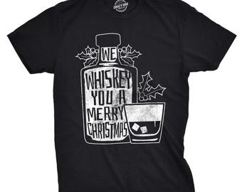 May include: A black short-sleeve t-shirt featuring a white graphic of a whiskey bottle and a glass with ice cubes. The bottle is labeled with the text WE WHISKEY YOU A MERRY CHRISTMAS and is decorated with holly leaves and berries on the sides.