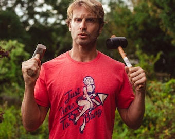 May include: A red short-sleeved t-shirt featuring a graphic print of a blonde woman sitting on a firework with the text Just Here To Bang. The shirt is worn by a person holding a metal hammer in one hand and a black rubber mallet in the other.
