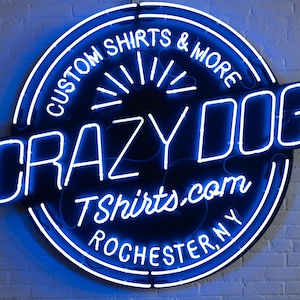 May include: A neon sign for Crazy Dog T-shirts & More, featuring a blue and white circular design with the text "Custom Shirts & More" and "Crazy Dog" in bold letters. The sign also includes the website address and the city, Rochester, NY.