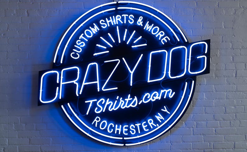 May include: A neon sign for Crazy Dog T-shirts & More, featuring a blue and white circular design with the text "Custom Shirts & More" and "Crazy Dog" in bold letters. The sign also includes the website address and the city, Rochester, NY.