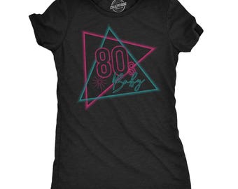 May include: A black short-sleeved t-shirt featuring a graphic design of two overlapping triangles in neon pink and cyan. The text 80's Baby is printed in the center in a stylized font. A small starburst icon is located to the left of the text.