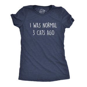 Puede incluir: Una camiseta de manga corta en color azul marino con textura jaspeada. La parte delantera presenta el texto blanco "I WAS NORMAL 3 CATS AGO" impreso en una fuente sencilla sin remates. La prenda tiene cuello redondo y un pequeño logo interior.