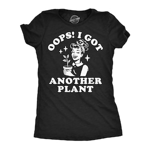May include: A black short-sleeved t-shirt featuring a white graphic print of a person holding a small potted plant. The text reads OOPS! I GOT ANOTHER PLANT in bold white lettering. The design includes small star accents and a retro-style illustration.