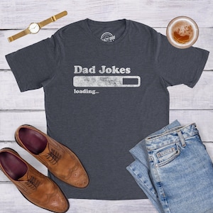 May include: A dark gray short-sleeve t-shirt featuring the white text Dad Jokes above a loading bar graphic and the word loading. The shirt is displayed with a gold-tone wristwatch, a pair of brown leather dress shoes, and blue denim jeans.
