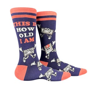 May include: A pair of navy blue crew socks with coral-colored cuffs, heels, and toes. The socks feature a repeating pattern of cartoon cassette tapes and floppy disks with arms and legs, and the text "THIS IS HOW OLD I AM" in coral.