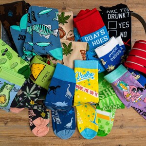 Puede incluir: Una colecci&oacute;n de calcetines coloridos con varios dise&ntilde;os y frases, incluyendo "Fitness Taco", "Vodka Made Me Do It", "Boats & Hoes", "She-nanigans" y "Crazy Dog".