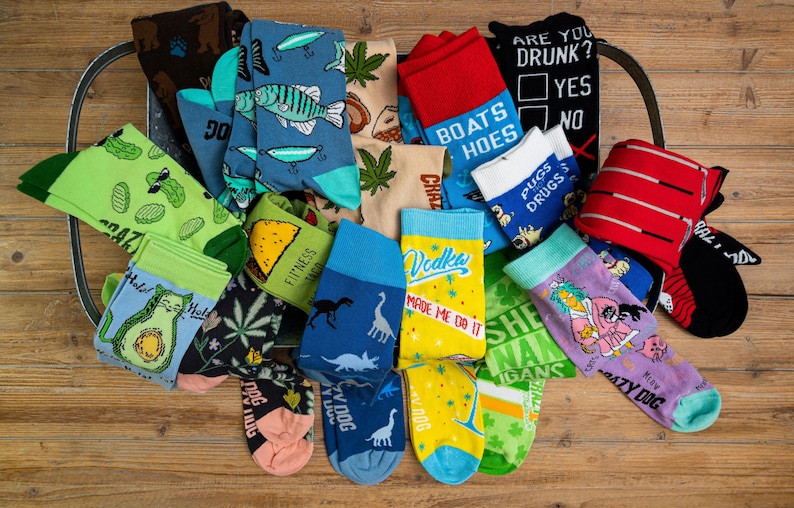 Puede incluir: Una colecci&oacute;n de calcetines coloridos con varios dise&ntilde;os y frases, incluyendo "Fitness Taco", "Vodka Made Me Do It", "Boats & Hoes", "She-nanigans" y "Crazy Dog".
