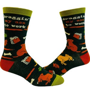 May include: A pair of dark green crew socks featuring a repeating pattern of orange dogs, white bones, red fire hydrants, and green paw prints. The text "draggin' my ass to work" is printed in white and orange. The socks have olive green cuffs and red heels.