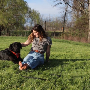 May include: A woman and a black dog are sitting in a grassy field. The dog wears a red bandana. The woman is wearing a gray t-shirt with the text "I LIKE DOGS AND MAYBE 3 PEOPLE". The woman is wearing blue jeans.