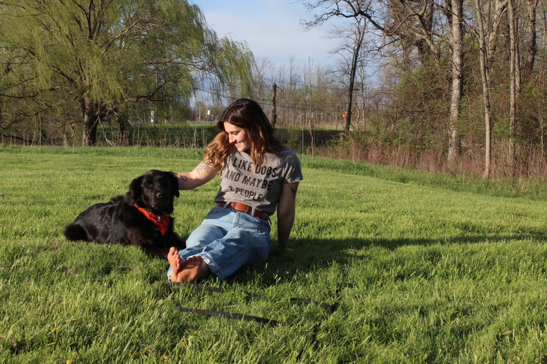 May include: A woman and a black dog are sitting in a grassy field. The dog wears a red bandana. The woman is wearing a gray t-shirt with the text "I LIKE DOGS AND MAYBE 3 PEOPLE". The woman is wearing blue jeans.