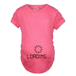 May include: A heathered pink short-sleeved t-shirt with a crew neckline. The front features a black graphic of a circular loading icon positioned above the text "LOADING..." in a digital, blocky font style centred on the lower torso area.