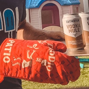 Op de afbeelding: Een rode ovenwant met de woorden "Patio Doddio" erop gedrukt. De want wordt gebruikt om twee blikjes Cutwater Vodka Mule vast te houden. De blikjes staan op een houten oppervlak.