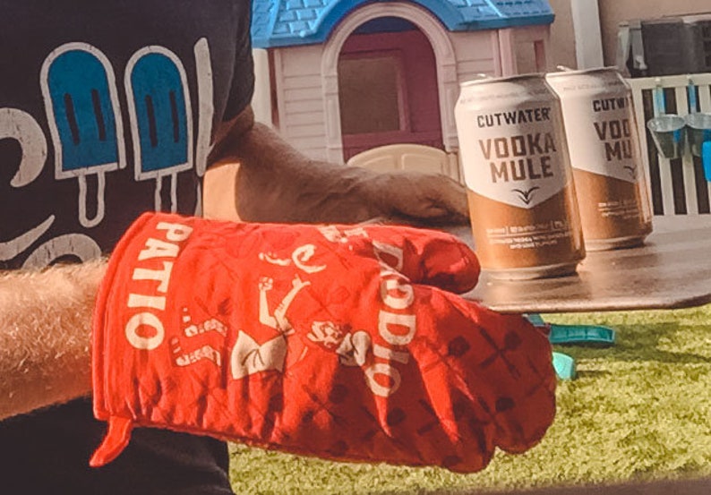 Op de afbeelding: Een rode ovenwant met de woorden "Patio Doddio" erop gedrukt. De want wordt gebruikt om twee blikjes Cutwater Vodka Mule vast te houden. De blikjes staan op een houten oppervlak.