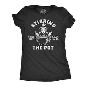 May include: A charcoal gray short-sleeve t-shirt featuring a white graphic of a witch with hands raised over a cauldron. The text reads STIRRING THE POT, SINCE 1692, and SALEM MASS. The design has a distressed, weathered appearance on the fabric.