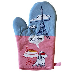 May include: Pink and blue oven mitt with a cartoon image of the Eiffel Tower, a dog, and a table with a baguette and a glass of wine. The text "Oui Oui" is in a speech bubble above the dog.