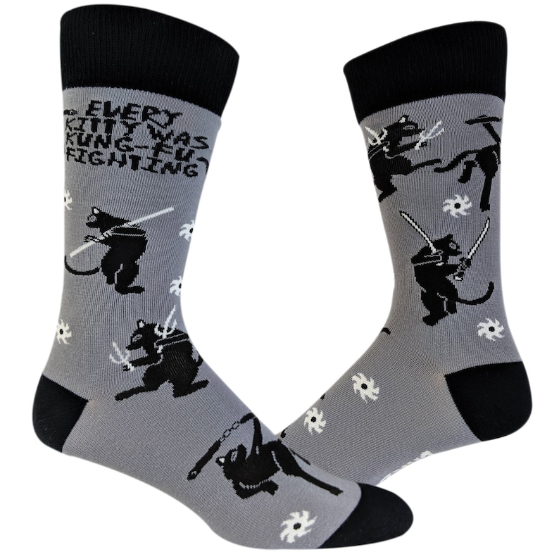 May include: A pair of gray socks with black and white illustrations of cats fighting with swords and throwing ninja stars. The socks have a black cuff and the text "EVERY KITTY WAS KUNG-FU FIGHTING" is printed on the front of the socks.