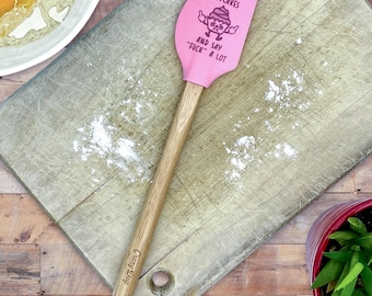 May include: A pink silicone spatula with a wooden handle. The spatula head features a graphic of a cupcake with arms and the text: I BAKE CUPCAKES AND SAY FUCK A LOT. The handle is engraved with the text Crazy Dog near the bottom end.