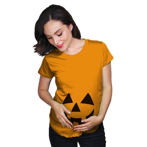 May include: A short-sleeved, crew-neck t-shirt in a vibrant orange colour. The front of the shirt features a large, black, graphic print of a jack-o'-lantern face, consisting of two triangular eyes, a triangular nose, and a jagged, toothy mouth.