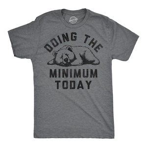 May include: A charcoal grey short-sleeve t-shirt featuring a black graphic of a sleeping bear. The text above the bear reads DOING THE and the text below reads MINIMUM TODAY in a bold, distressed font. The shirt has a crew neckline.