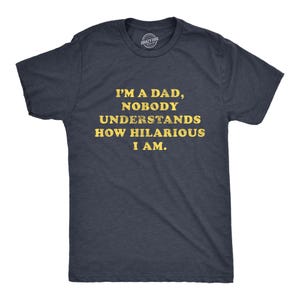 May include: A dark navy blue short-sleeved t-shirt featuring the yellow distressed text I'M A DAD, NOBODY UNDERSTANDS HOW HILARIOUS I AM printed on the front. The shirt has a crew neckline and a standard fit, with a small circular logo at the neck.