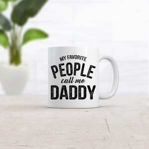 Puede incluir: Una taza de cerámica blanca con el texto negro MY FAVORITE PEOPLE call me DADDY impreso en el frente. La taza tiene una forma cilíndrica estándar con un asa curva en el lado derecho y un acabado suave y brillante en todo el exterior.