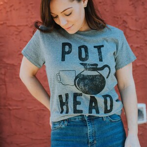 May include: A heather gray short-sleeve t-shirt featuring a black graphic print of a coffee pot and a mug with the text POT HEAD centered on the front. The design has a distressed, textured appearance and is printed on a standard crew neck shirt.