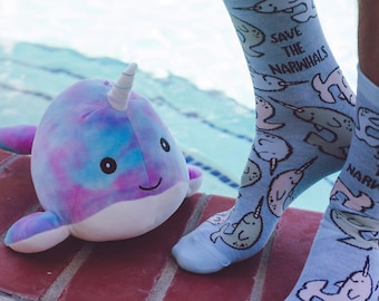 May include: A plush narwhal toy with a tie-dye blue and purple pattern and a white horn, alongside light blue socks featuring a repeating narwhal print and the text "SAVE THE NARWHALS" in black lettering, positioned on a red brick surface.