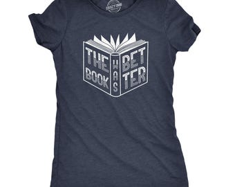 May include: A dark navy blue short-sleeve t-shirt featuring a white graphic of an open book. The text on the book reads THE BOOK WAS BETTER, with the word WAS printed vertically along the spine. The shirt has a crew neckline and a heathered texture.