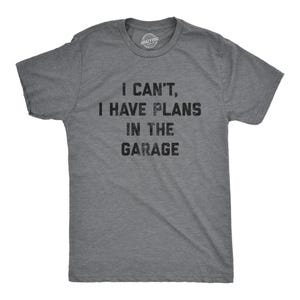 May include: A heather grey short-sleeve t-shirt featuring the black distressed text I CAN'T, I HAVE PLANS IN THE GARAGE printed on the front. The shirt has a crew neckline and a standard fit, showcasing a casual style with a clear, bold graphic.