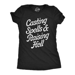 May include: A black short-sleeved t-shirt featuring white distressed-style text printed on the front that reads Casting Spells & Raising Hell. The shirt has a crew neckline and a standard fit, with a small brand logo visible inside the neck area.