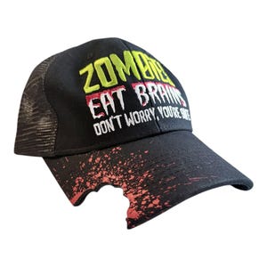 Puede incluir: Gorra de camionero negra con el texto "ZOMBIE EAT BRAINS DON'T WORRY, YOU'RE SAFE" en amarillo, rojo y blanco. La gorra tiene una parte trasera de malla y un diseño de salpicaduras de sangre en la visera.