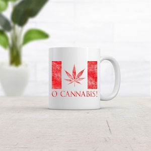 May include: A white ceramic mug featuring a red graphic design that mimics the Canadian flag, with a cannabis leaf centered in the middle. Below the flag graphic, the text O CANNABIS! is printed in a bold, red, distressed-style font.