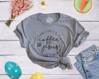 Op de afbeelding: Een grijs gemêleerd T-shirt met korte mouwen en een zwarte cirkelvormige opdruk met de tekst, Today is going to be a coffee and jesus kind of day. Het shirt is onderaan geknoopt en ligt tussen roze bloemen, een madeliefje, een wortelvormig koekje en twee gestippelde paaseieren.