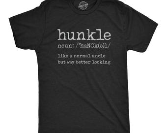 Puede incluir: Una camiseta negra de manga corta con texto blanco que dice: hunkle noun: /'huNGk(ə)l/ like a normal uncle but way better looking. La prenda tiene un cuello redondo y un pequeño logotipo circular impreso en el interior del cuello.