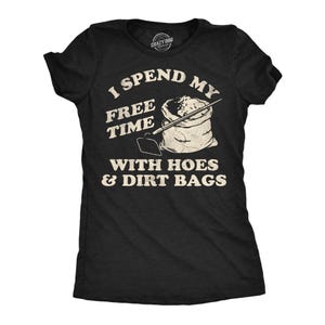 May include: A black short-sleeved t-shirt featuring a graphic print in off-white. The design includes a drawing of a garden hoe and a sack of soil, with the text: I SPEND MY FREE TIME WITH HOES & DIRT BAGS. The shirt has a crew neckline.