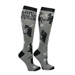 May include: A pair of gray knee-high socks with a black cat design. The socks feature a repeating pattern of black cats holding swords and throwing stars. The text "EVERY KITTY WAS KUNG-FU FIGHTING" is printed on the socks.