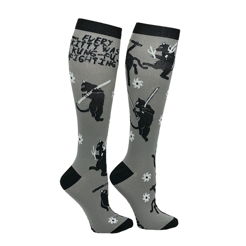 May include: A pair of gray knee-high socks with a black cat design. The socks feature a repeating pattern of black cats holding swords and throwing stars. The text "EVERY KITTY WAS KUNG-FU FIGHTING" is printed on the socks.