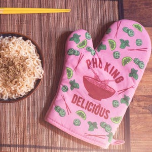 May include: A pink oven mitt featuring a repeating pattern of lime wedges, herbs, and chopsticks. The center displays a graphic of a bowl of noodles with the text PHO KING DELICIOUS printed in dark red letters across the fabric surface.